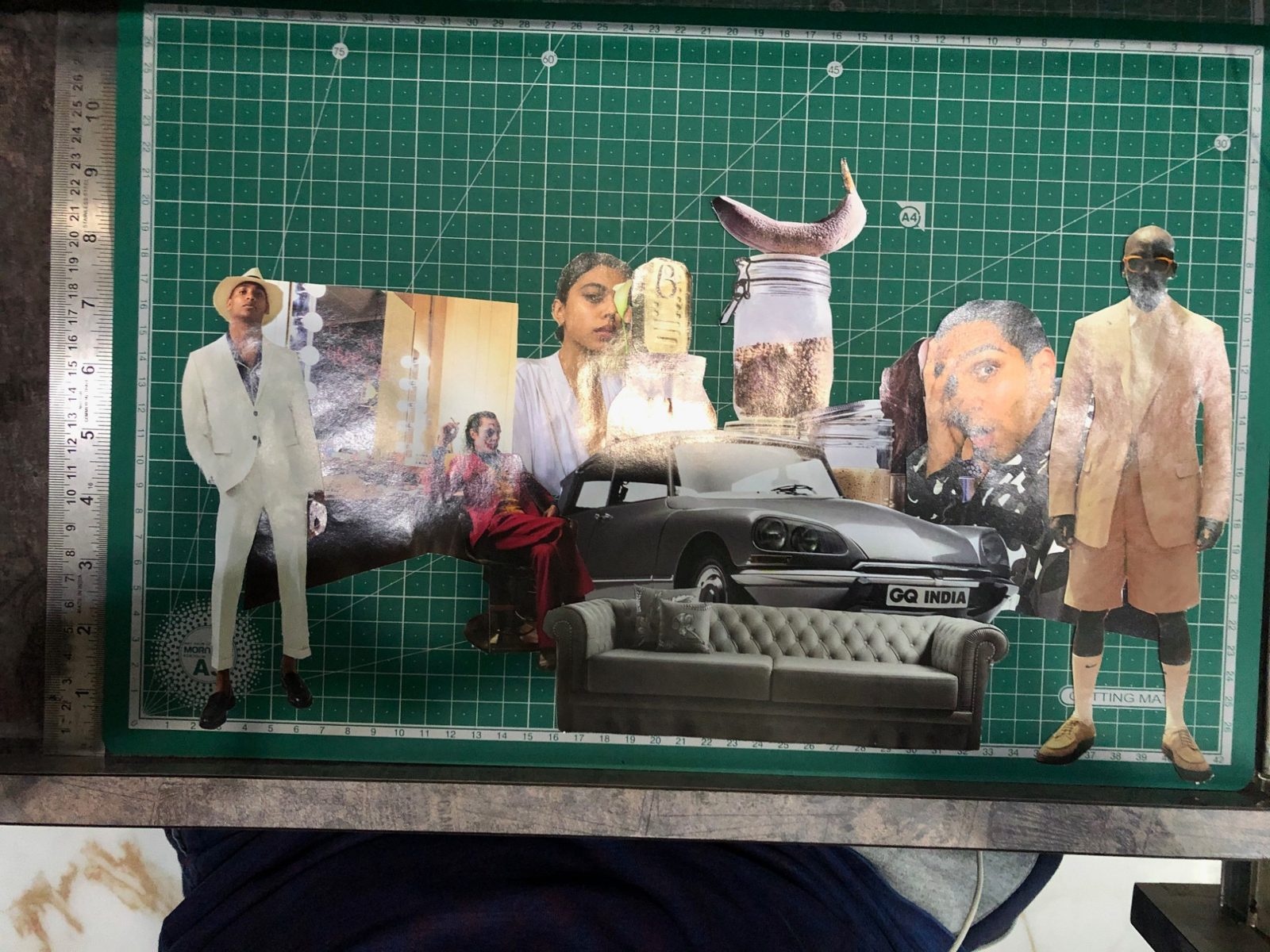 An arrangement in progress on the cutting mat: a white-suited figure, a grey vintage car badged GQ India, a tufted sofa, a banana and assorted cut-out portraits